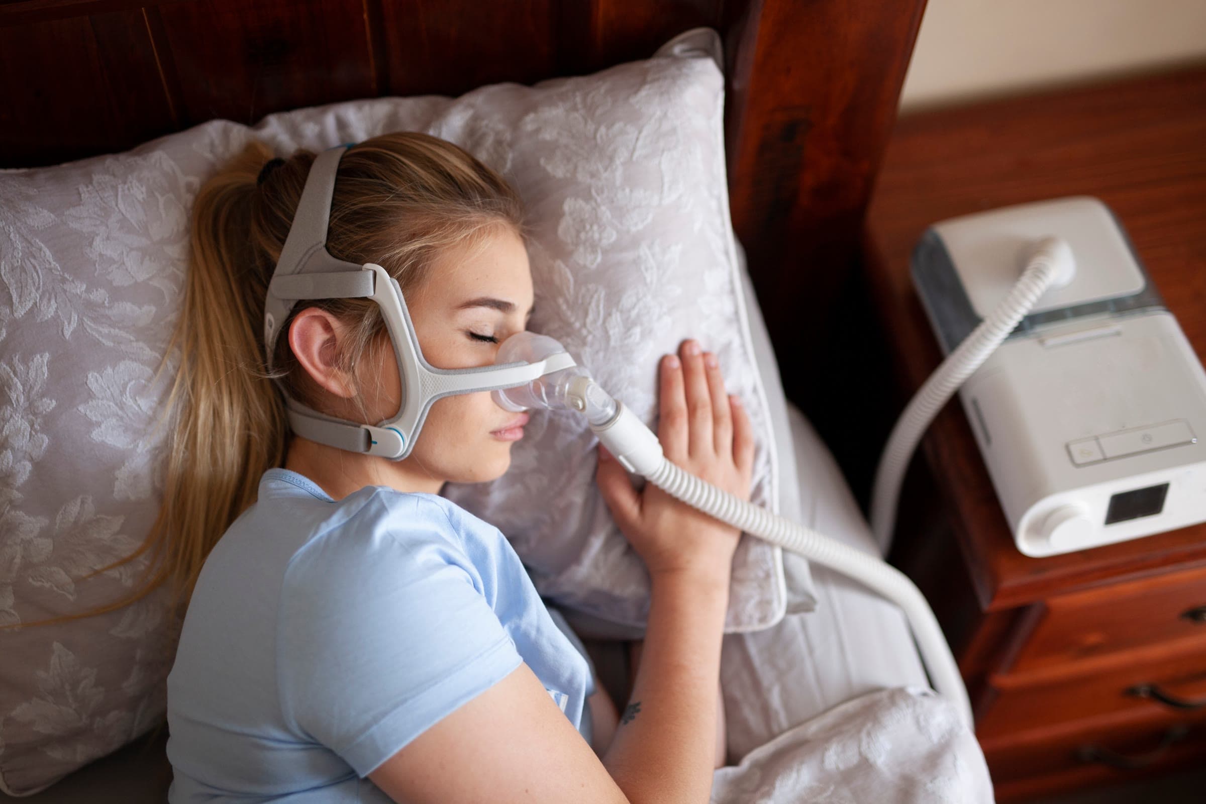 A woman asleep on her side wearing a CPAP nasal mask, with the machine on the bedside table.